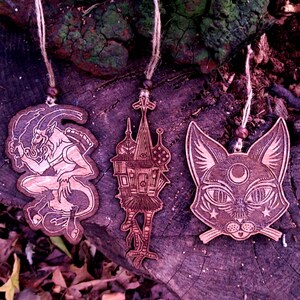 May include: Three wooden ornaments hanging from twine. The ornaments are laser cut and feature a black cat with a crescent moon on its forehead, a house with a bird-like creature, and a horned creature with a tail.