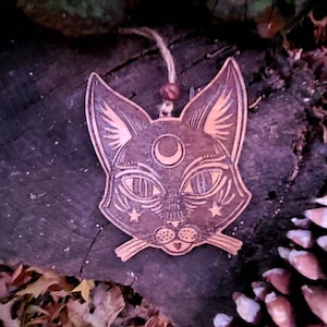 May include: A wooden cat head ornament with a crescent moon and stars on its face. The ornament is painted in shades of brown and black and has a rustic, vintage look.
