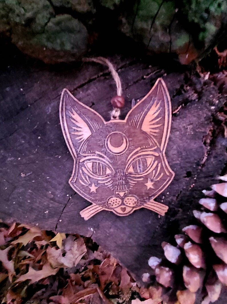 May include: A wooden cat head ornament with a crescent moon and stars on its face. The ornament is painted in shades of brown and black and has a rustic, vintage look.