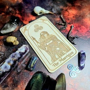 K&ouml;nnte beinhalten: Eine Tarotkarte aus Holz mit dem Text "The Morrigan" und einer Illustration einer Frau in einem Umhang, umgeben von Kristallen, einem Vogelsch&auml;del und anderen mystischen Gegenst&auml;nden. Die Karte zeigt keltische Knoten und Fledermaussymbole.