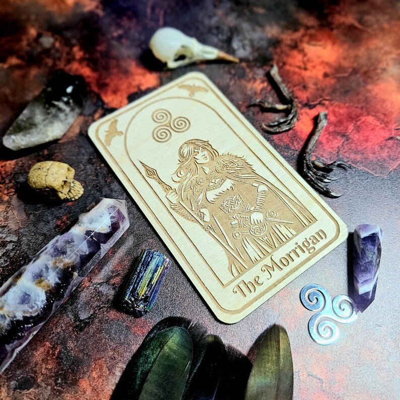 K&ouml;nnte beinhalten: Eine Tarotkarte aus Holz mit dem Text "The Morrigan" und einer Illustration einer Frau in einem Umhang, umgeben von Kristallen, einem Vogelsch&auml;del und anderen mystischen Gegenst&auml;nden. Die Karte zeigt keltische Knoten und Fledermaussymbole.