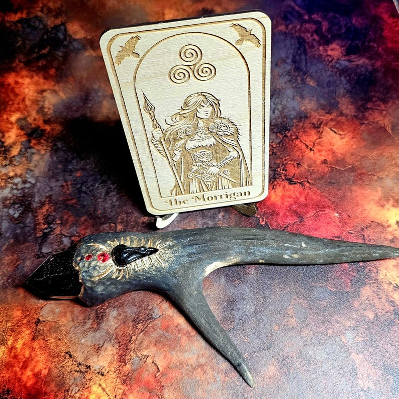 K&ouml;nnte beinhalten: Eine Tarotkarte aus Holz mit dem Text "The Morrigan" und einem geschnitzten Bild einer Frau mit einem Speer. Darunter befindet sich ein geschnitzter Holzhahn mit roten Augen und einem schwarzen Schnabel.