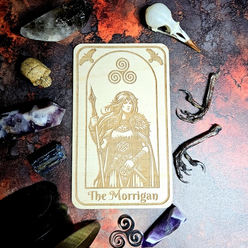 K&ouml;nnte beinhalten: Eine Tarotkarte aus Holz mit einer gravierten Illustration einer Frau, die einen Speer h&auml;lt, mit dem Text "The Morrigan". Die Karte ist von Kristallen, einem Vogelsch&auml;del und anderen mystischen Gegenst&auml;nden umgeben. Die Karte hat eine hellbraune Farbe.