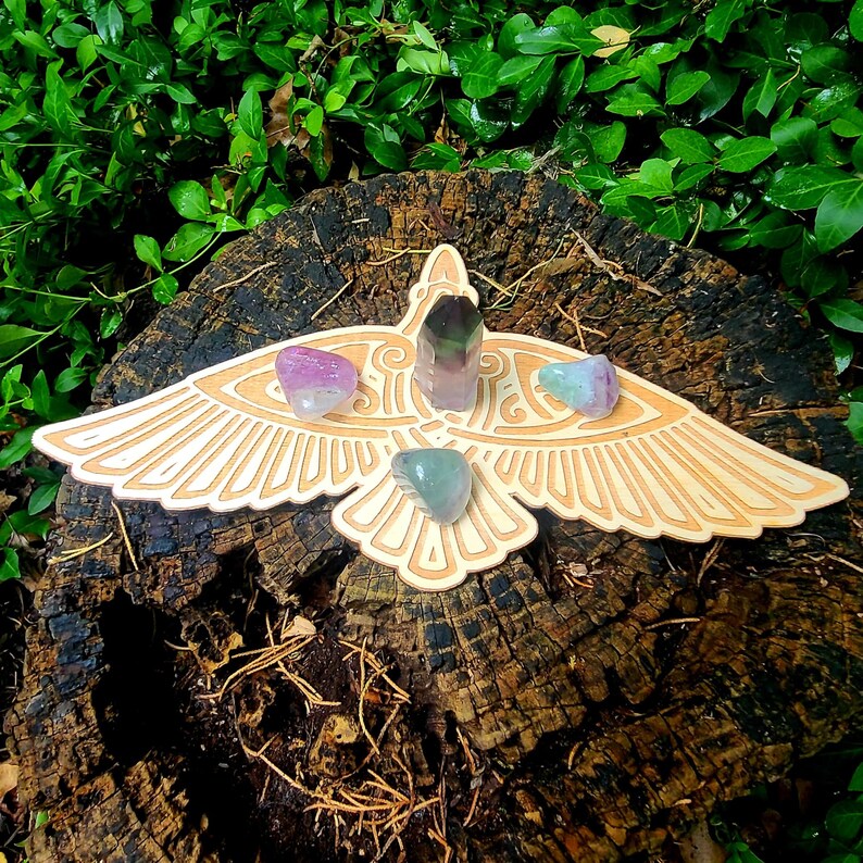 May include: A wooden bird-shaped crystal grid with four crystals placed on it. The crystals are green, purple, and pink. The grid is on a tree stump.