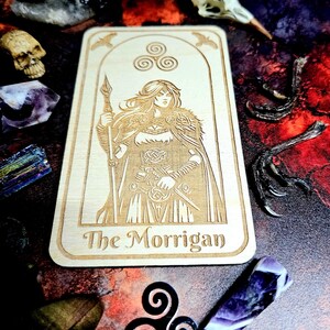 K&ouml;nnte beinhalten: Eine hellbraune Tarotkarte aus Holz mit einer Gravur einer Frau, die einen Speer und ein Schwert h&auml;lt, mit dem Text "The Morrigan". Die Karte ist von Kristallen, einem Sch&auml;del und Vogelkrallen umgeben. Das Thema ist mystisch.