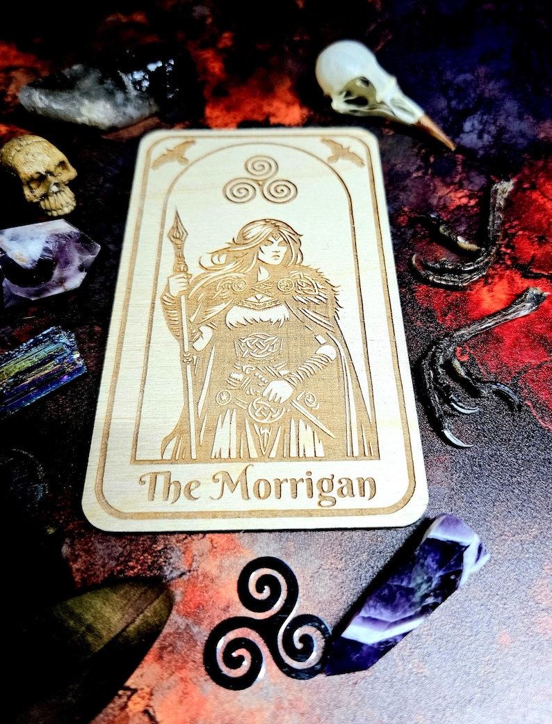 K&ouml;nnte beinhalten: Eine hellbraune Tarotkarte aus Holz mit einer Gravur einer Frau, die einen Speer und ein Schwert h&auml;lt, mit dem Text "The Morrigan". Die Karte ist von Kristallen, einem Sch&auml;del und Vogelkrallen umgeben. Das Thema ist mystisch.