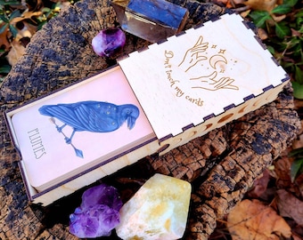 May include: A wooden box with a light wood finish and a black raven illustration on the front. The box has a lid with the text "Don't touch my cards" engraved on it. The box is sitting on a tree stump with some crystals around it.