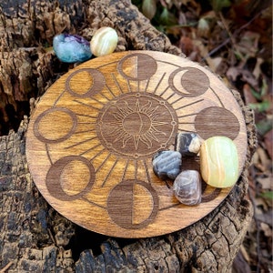 May include: A wooden moon phase board with a sun and moon design. The board is surrounded by various crystals, including clear, white, and black.