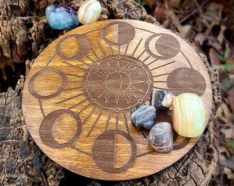 May include: A wooden moon phase board with a sun and moon design. The board is surrounded by various crystals, including clear, white, and black.