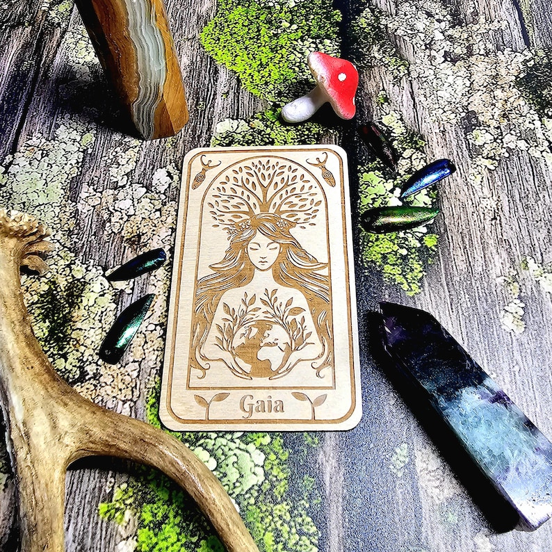 May include: A wooden rectangular card with the word "Gaia" at the bottom, featuring an engraved illustration of a woman holding the Earth, surrounded by a tree and leaves. The image also includes decorative crystals, a mushroom, and iridescent beetle wings.