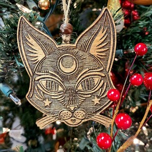 May include: A wooden cat ornament with a crescent moon and stars carved into its face. The ornament is hanging on a Christmas tree with red berries and green pine needles.