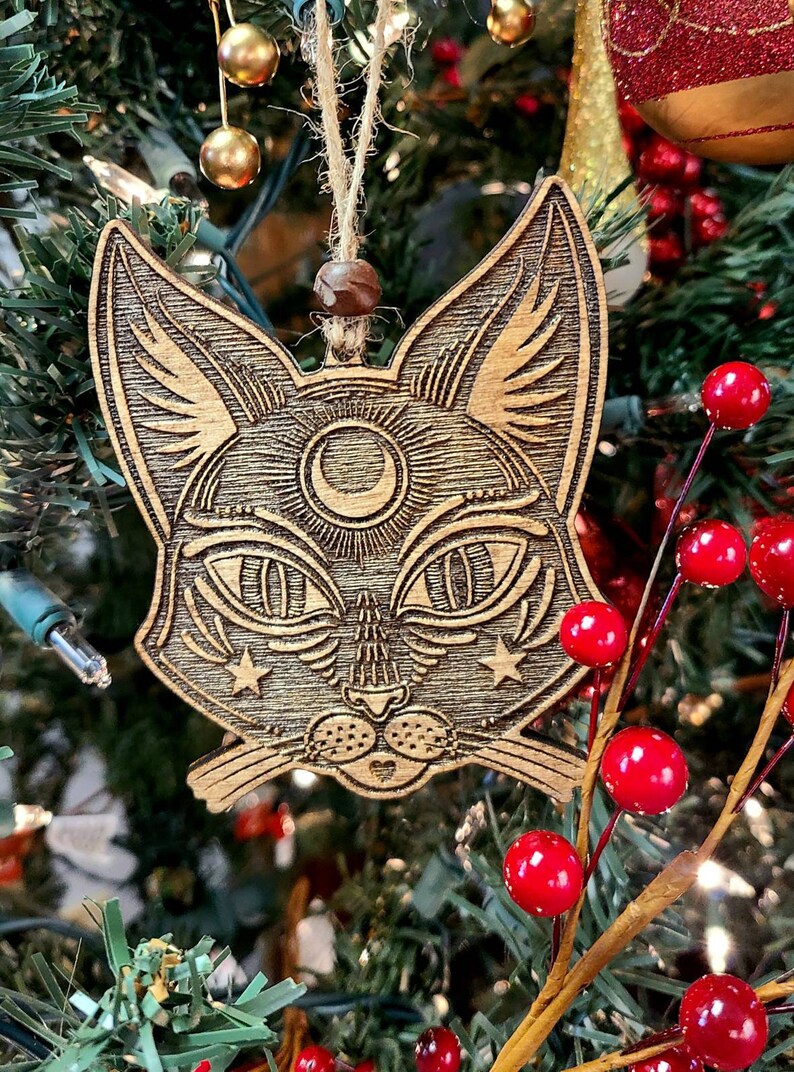May include: A wooden cat ornament with a crescent moon and stars carved into its face. The ornament is hanging on a Christmas tree with red berries and green pine needles.
