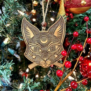 May include: A wooden cat ornament with a crescent moon and stars on its face. The ornament is hanging on a Christmas tree with red berries.