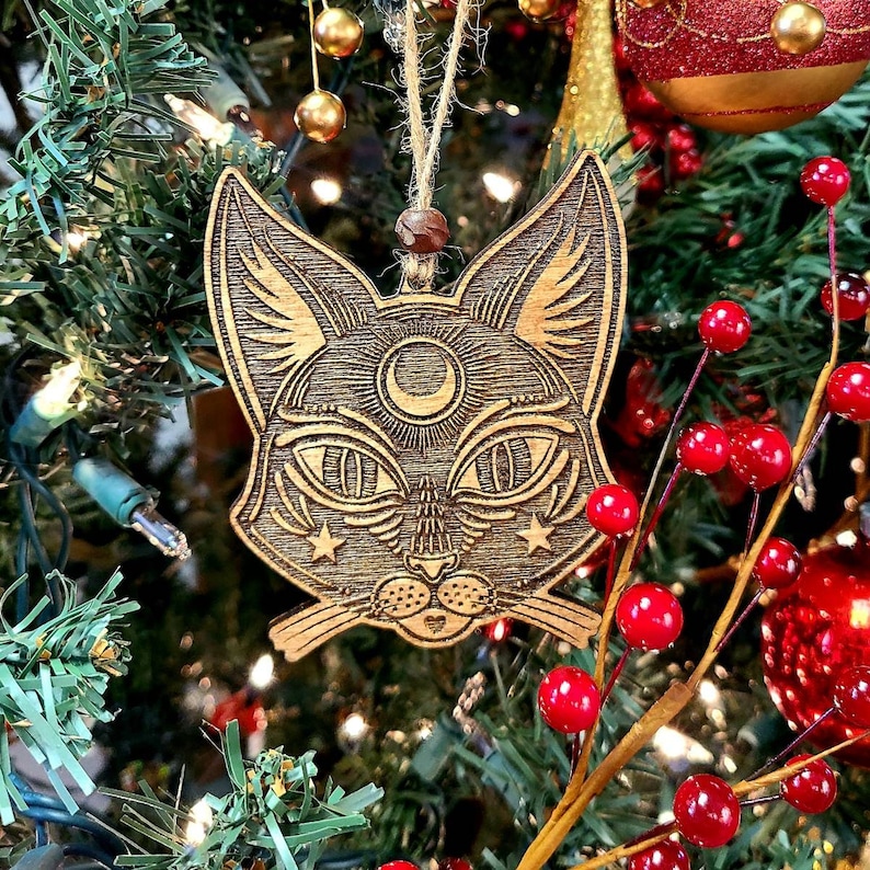 May include: A wooden cat ornament with a crescent moon and stars on its face. The ornament is hanging on a Christmas tree with red berries.
