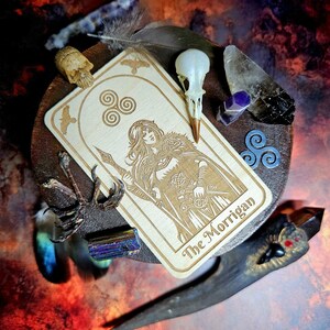 K&ouml;nnte beinhalten: Eine Tarotkarte aus Holz mit einer Gravur von "The Morrigan", umgeben von Federn, Kristallen und einem Vogelsch&auml;del. Die Farbgebung umfasst Braun-, Beige- und Blaunuancen sowie Violett. Das Thema ist mystisch und spirituell.