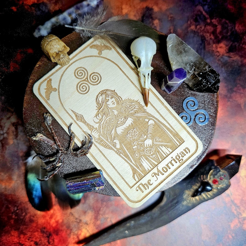 K&ouml;nnte beinhalten: Eine Tarotkarte aus Holz mit einer Gravur von "The Morrigan", umgeben von Federn, Kristallen und einem Vogelsch&auml;del. Die Farbgebung umfasst Braun-, Beige- und Blaunuancen sowie Violett. Das Thema ist mystisch und spirituell.