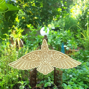 May include: A wooden cutout of a stylized bird with intricate patterns. The bird is light brown and has a detailed design on its wings and body. The bird is placed in a garden setting with green plants.
