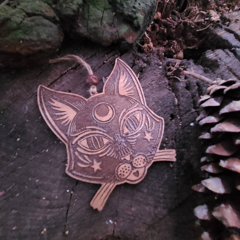 May include: A wooden cat ornament with a crescent moon and stars carved into its face. The ornament is painted in a dark brown color and has a rustic look.