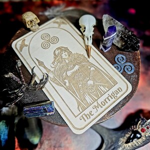 K&ouml;nnte beinhalten: Eine Holz-Tarotkarte mit einer Gravur einer Frau mit Speer, mit dem Text "The Morrigan". Die Karte ist umgeben von Kristallen, einem Vogelsch&auml;del und anderen mystischen Objekten.