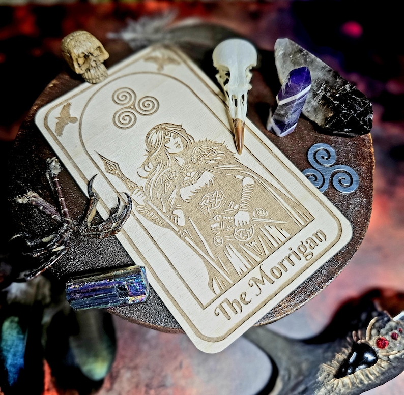 K&ouml;nnte beinhalten: Eine Holz-Tarotkarte mit einer Gravur einer Frau mit Speer, mit dem Text "The Morrigan". Die Karte ist umgeben von Kristallen, einem Vogelsch&auml;del und anderen mystischen Objekten.