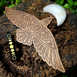 May include: A wooden carving of a stylized raven with intricate details, set against a backdrop of a tree stump and a white crescent moon. The raven's wings are spread wide, and its body is adorned with Celtic knotwork.