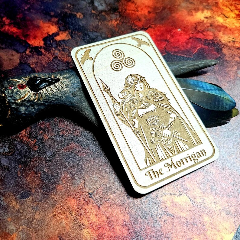 K&ouml;nnte beinhalten: Helle Holz-Tarotkarte mit graviertem Bild einer Frau mit Speer und dem Text "The Morrigan" unten. Die Karte hat einen dekorativen Rahmen mit drei Spiralen und zwei V&ouml;geln. Das Design ist hellbraun.