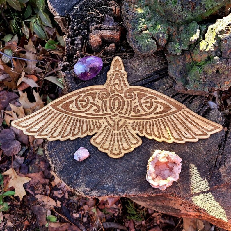 May include: A wooden carving of a stylized raven with intricate details. The raven is spread-winged and has a Celtic knot design on its body. The carving is on a natural wood background with small pink and purple crystals.