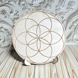 Peut inclure: Un objet d&eacute;coratif rond en bois clair avec un motif g&eacute;om&eacute;trique. Le motif pr&eacute;sente des cercles qui s'entrecroisent, cr&eacute;ant un motif floral. La pi&egrave;ce est soutenue par un petit support en bois.