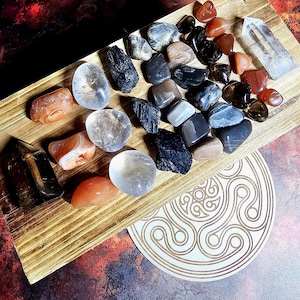 Puede incluir: Una colección de piedras preciosas pulidas de diversas formas y colores, incluyendo cuarzo transparente, turmalina negra y cornalina, dispuestas sobre una tabla de madera. Las piedras se exhiben junto con una punta de cristal de cuarzo transparente y un posavasos decorativo de madera.