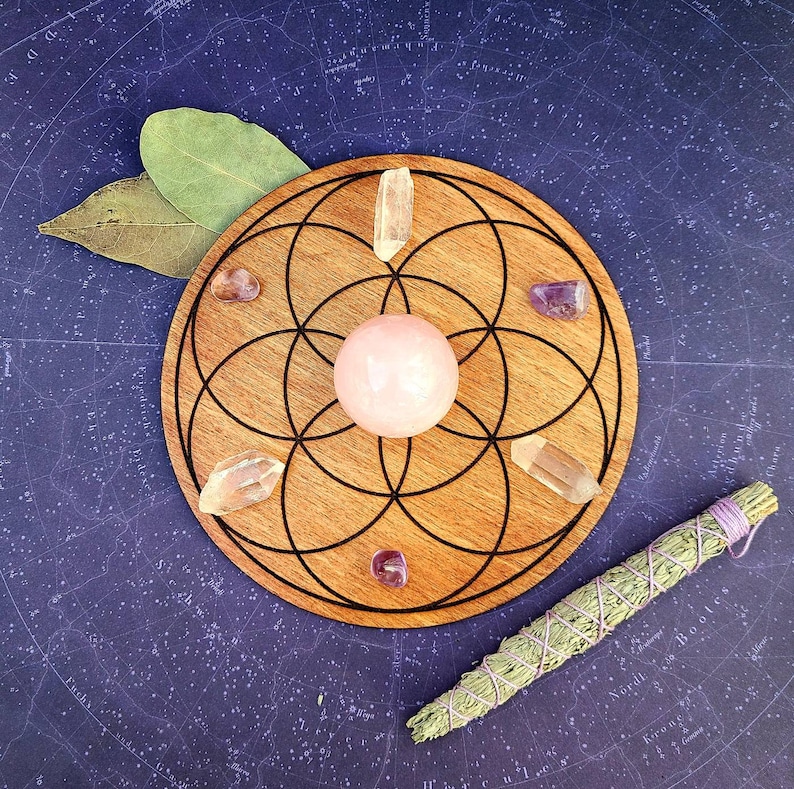 Peut inclure: Un support en bois pour cristaux avec un motif g&eacute;om&eacute;trique noir, contenant une sph&egrave;re rose, des cristaux de quartz clair et des pierres d'am&eacute;thyste. Un paquet de sauge et des feuilles de laurier sont &eacute;galement pr&eacute;sents. Le fond est une surface bleu fonc&eacute; avec un motif d'&eacute;toiles.