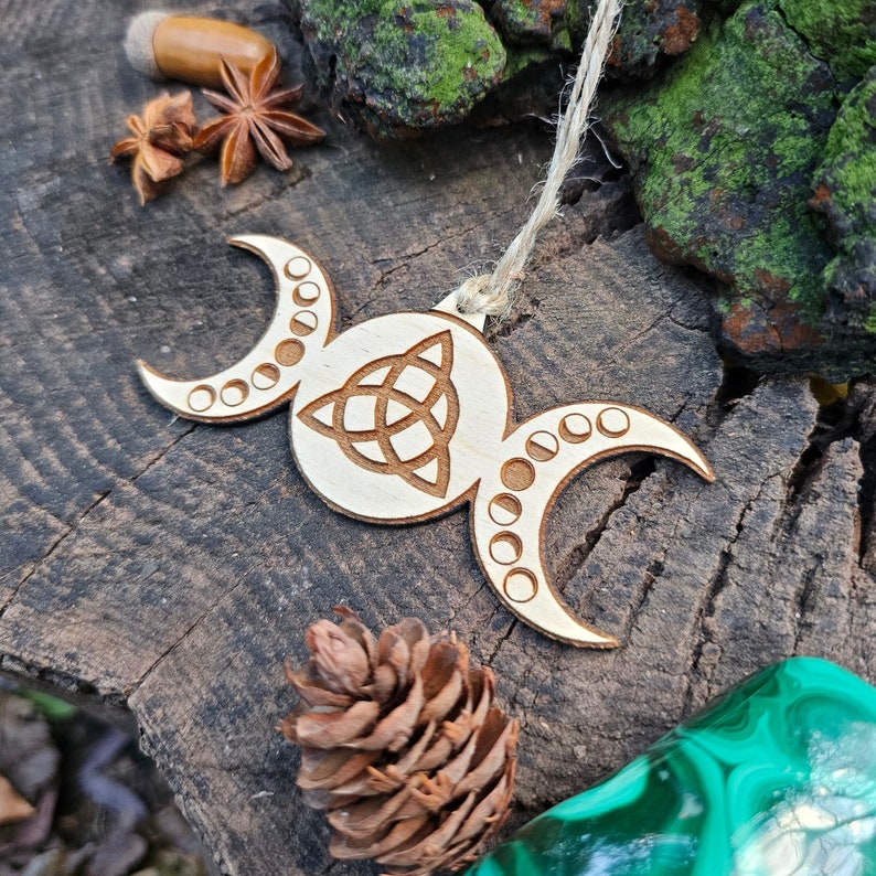 A lasercut and engraved wooden ornament hanging from thin twine. The ornament is in the shape of a full moon flanked by crescent moons, with moon phases and the Celtic triquetra symbols engraved into them.