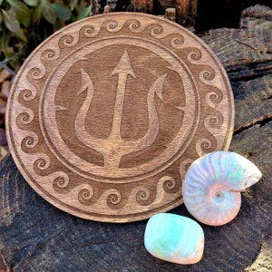 May include: A wooden coaster with a carved trident design surrounded by a wave pattern. The coaster is on a wooden surface with two iridescent stones.
