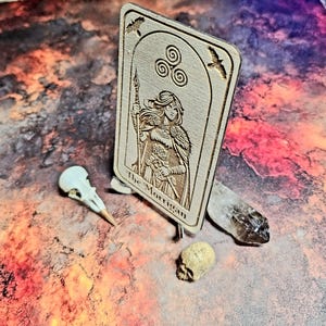 K&ouml;nnte beinhalten: Holz-Tarotkarte mit eingraviertem Bild einer Frau, die einen Speer h&auml;lt, mit der Aufschrift "The Morrigan". Die Karte wird von einem Vogelsch&auml;del, einem kleinen Sch&auml;del und einem Kristall begleitet.