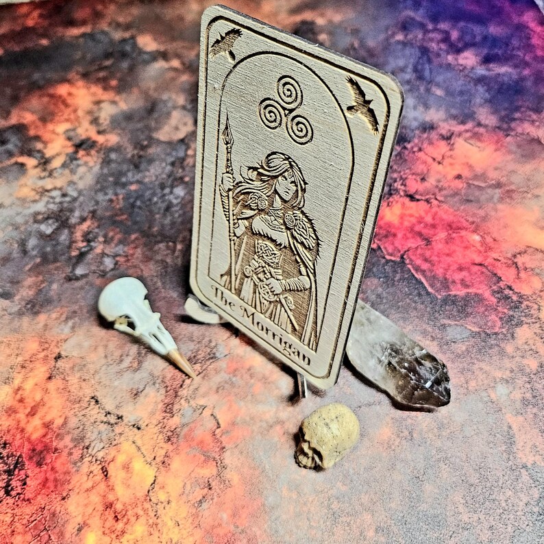 K&ouml;nnte beinhalten: Holz-Tarotkarte mit eingraviertem Bild einer Frau, die einen Speer h&auml;lt, mit der Aufschrift "The Morrigan". Die Karte wird von einem Vogelsch&auml;del, einem kleinen Sch&auml;del und einem Kristall begleitet.