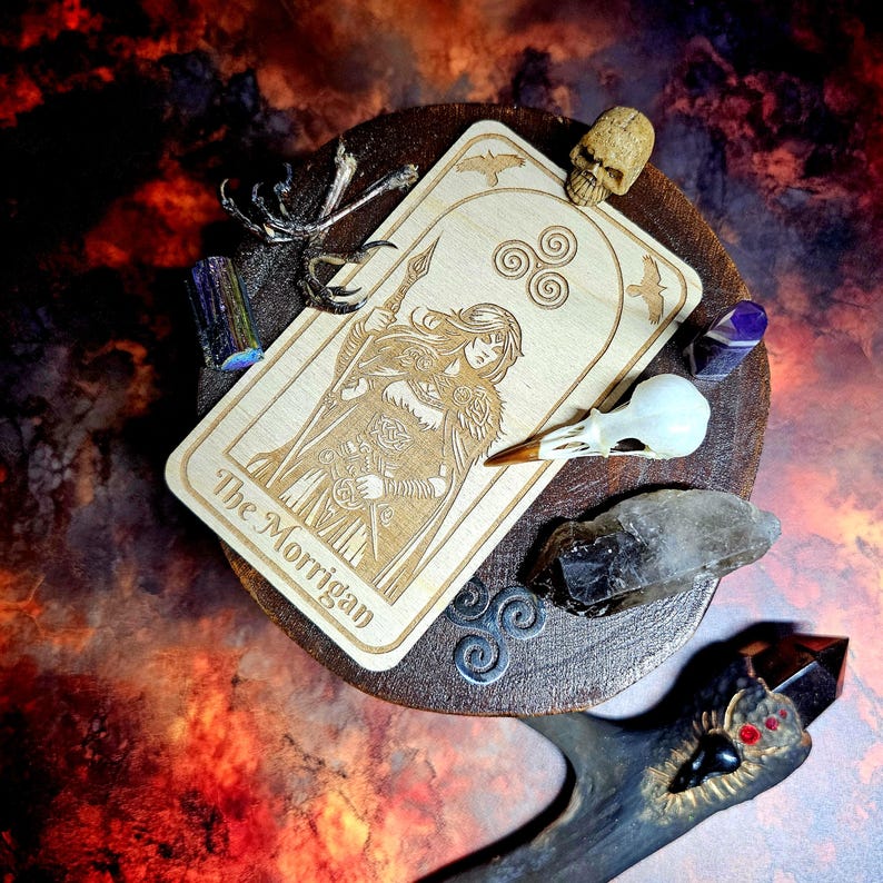K&ouml;nnte beinhalten: Eine Tarotkarte aus Holz mit dem Text "The Morrigan" und einer eingravierten Illustration einer Frau, die einen Speer h&auml;lt. Die Karte ist von Kristallen, einem Sch&auml;del, einem Vogelsch&auml;del und einem dekorativen Metallst&uuml;ck auf einem Holzsockel umgeben.