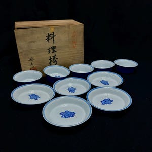 以下が含まれることがあります： 木箱に入った日本の陶器の食器セット。食器は、内側が白で青い花の模様と青い縁があります。木箱には日本語の文字が書かれています。