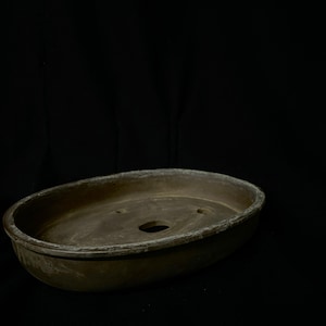 May include: A weathered, oval-shaped bonsai pot in a muted brown hue. The pot features three drainage holes and a slightly textured rim. The overall design suggests a rustic, natural aesthetic, suitable for bonsai cultivation.