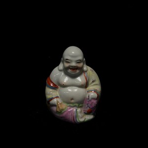 Peut inclure: Petite figurine de Bouddha en céramique blanche souriante. Le Bouddha est assis et porte une robe avec des accents rouges, jaunes et roses. La figurine a une finition brillante et est posée sur un fond noir.