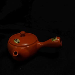 以下が含まれることがあります： 赤色の取っ手と蓋が付いた陶器のティーポット。 ティーポットには注ぎ口と小さな丸い蓋が付いています。 日本語のテキストが入ったステッカーが側面にあります。 ティーポットは黒い背景にあります。