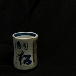 以下が含まれることがあります： 青と茶色のアクセントが付いた、小さな白いセラミックカップ。カップには青色の日本語の文字と青色の縁があります。カップは円筒形で、黒い表面に置かれています。