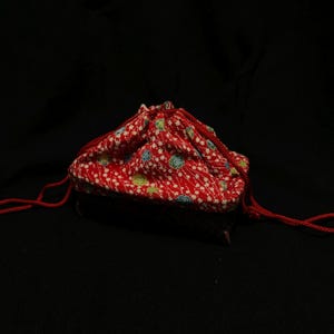 Könnte beinhalten: Ein kleiner Beutel mit Kordelzug aus einem leuchtend roten Stoff, der mit einem dichten Muster aus weißen Blumenzweigen sowie kleinen blauen und grünen kreisförmigen Akzenten verziert ist. Der Beutel wird oben mit zwei roten Kordeln zugezogen.