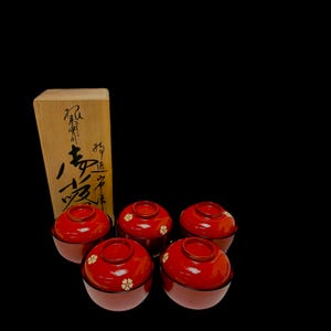 Peut inclure: Un ensemble de cinq bols à soupe japonais laqués rouges et bruns avec couvercles, chacun orné d'un motif floral blanc. Les bols sont disposés devant une boîte en bois avec calligraphie japonaise.
