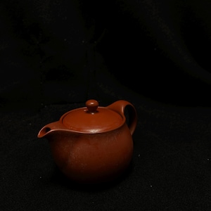 Puede incluir: Una pequeña tetera redonda de color marrón rojizo con una tapa a juego y un asa curvada. La tetera tiene un pico para verter y un pequeño pomo en la parte superior de la tapa. La tetera es de cerámica.