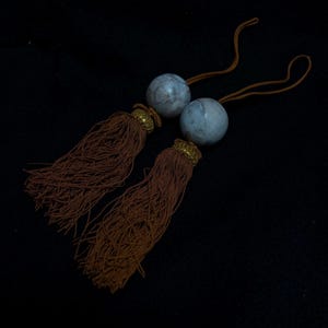 Puede incluir: Dos borlas decorativas con flecos marrón claro y detalles dorados. Cada borla está rematada con una esfera azul claro veteada y una tapa dorada. Las borlas están conectadas por un fino cordón marrón.