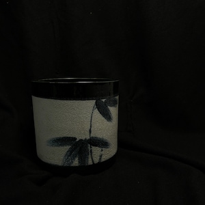 Op de afbeelding: Een cilindrische keramische beker met een getextureerde, gebroken witte buitenkant en een zwarte rand. De beker heeft een donkerblauw bloemmotief. De achtergrond is effen zwart.