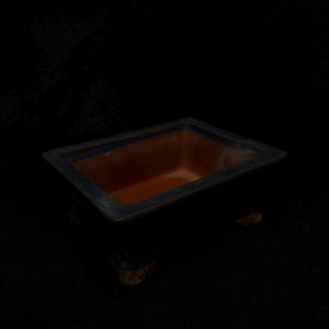 May include: A rectangular bonsai pot with a dark blue exterior and a reddish-brown interior. The pot has a slightly raised rim and sits on small, decorative feet. The overall design is simple and elegant, suitable for showcasing a small tree or plant.