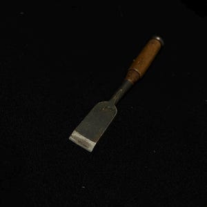 Pode incluir: Um pequeno cinzel antigo com cabo de madeira e lâmina de metal. A lâmina é retangular e parece desgastada, com uma borda ligeiramente arredondada. O cabo é marrom escuro e o metal é prateado fosco.