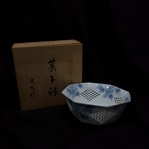 Puede incluir: Un cuenco de cerámica blanco y azul con un diseño floral y una caja de madera. El cuenco tiene una forma geométrica con lados perforados. El patrón floral azul decora el borde y los lados del cuenco. La caja de madera tiene texto japonés.