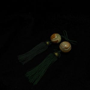 May include: A pair of decorative tassels featuring smooth, round, banded agate beads in shades of brown, cream, and tan. Each bead is topped with a small, textured, dark metallic cap and attached to a long, dark green, fine-threaded silk tassel.