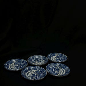 May include: Five small, round, blue and white porcelain plates with floral patterns. The plates have scalloped edges and a gold trim. The plates are set against a black backdrop.